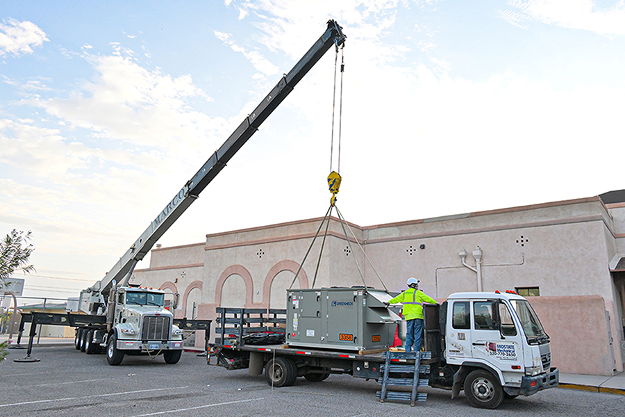 A truck with a crane picks up an A/C unit from another truck while a man in a yellow safety vest watches