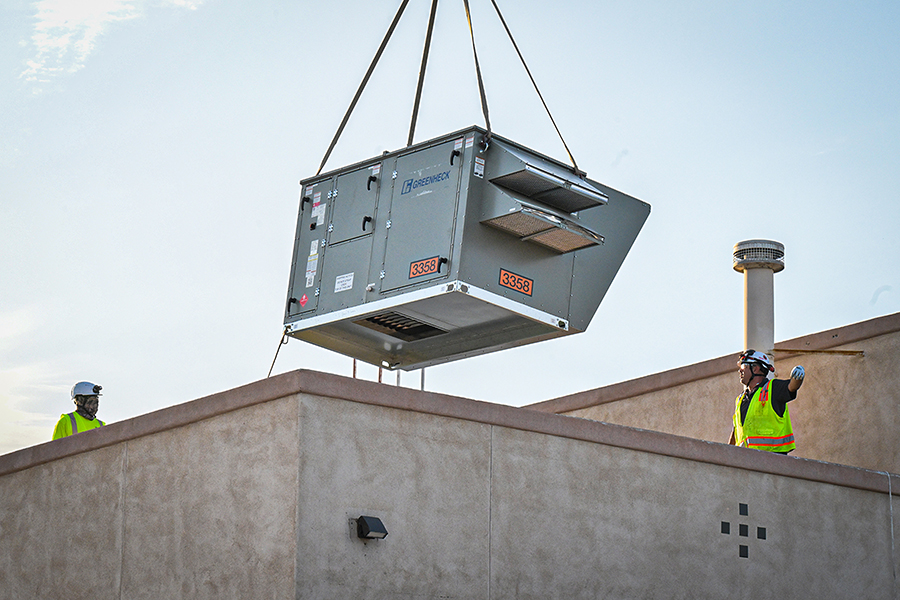 Two men in yellow safety vest and white hard hats oversee an A/C unit being lowered onto the roof