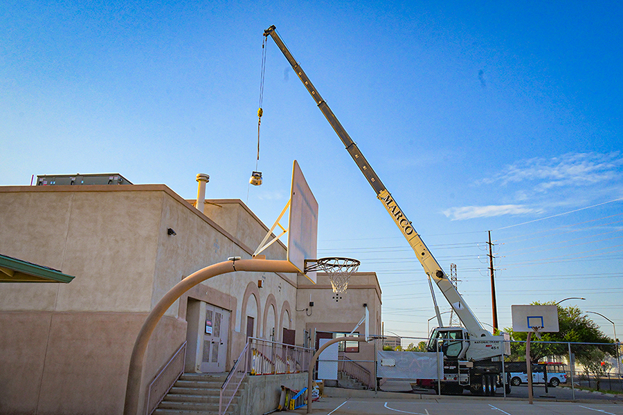 A basketball hoop in the foreground, with a crane lifting an A/C unit onto a roof in the background