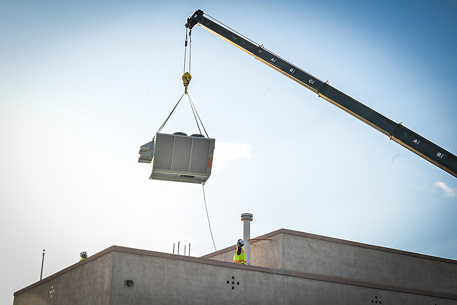 A man in a yellow safety vest supervises an A/C unit being lowered on the roof