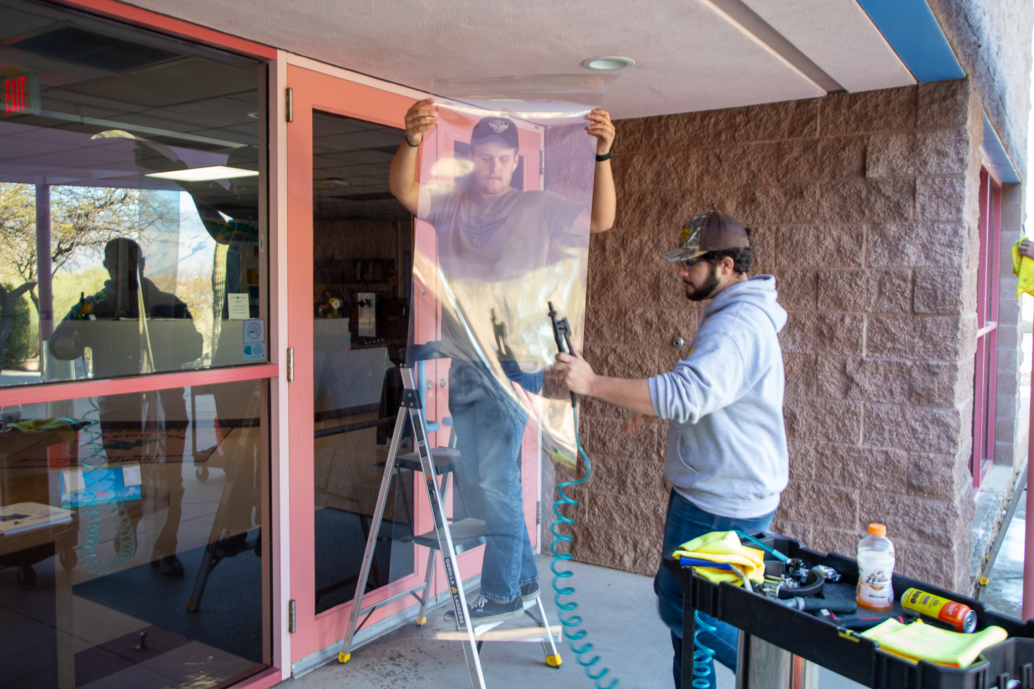 A man stands on a step ladder holding up a security film while another man adjusts it