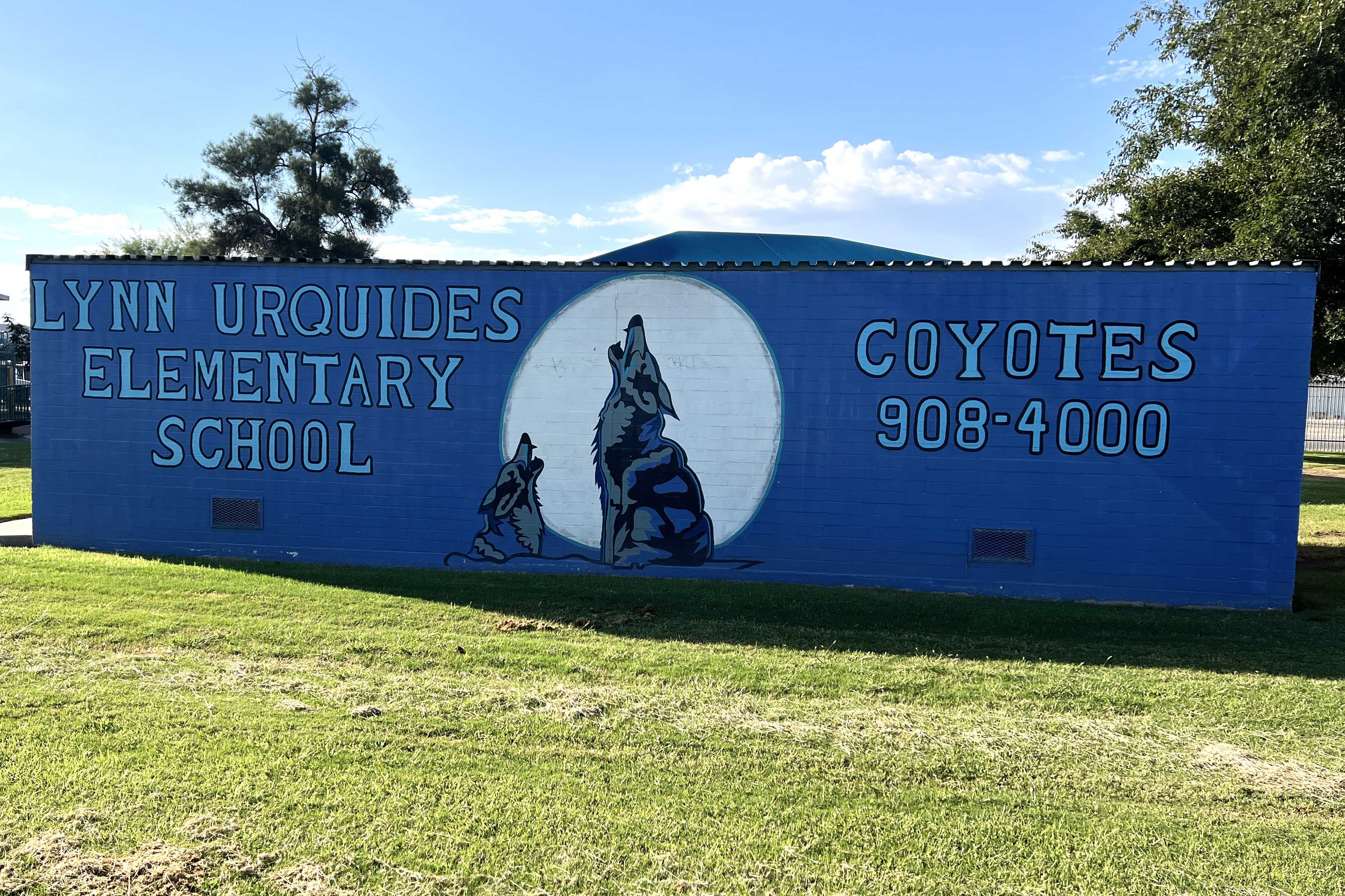 A blue brick wall with a mural of two coyotes howling at the moon reading Lynn Urquides Elementary School Coyotes 908-4000