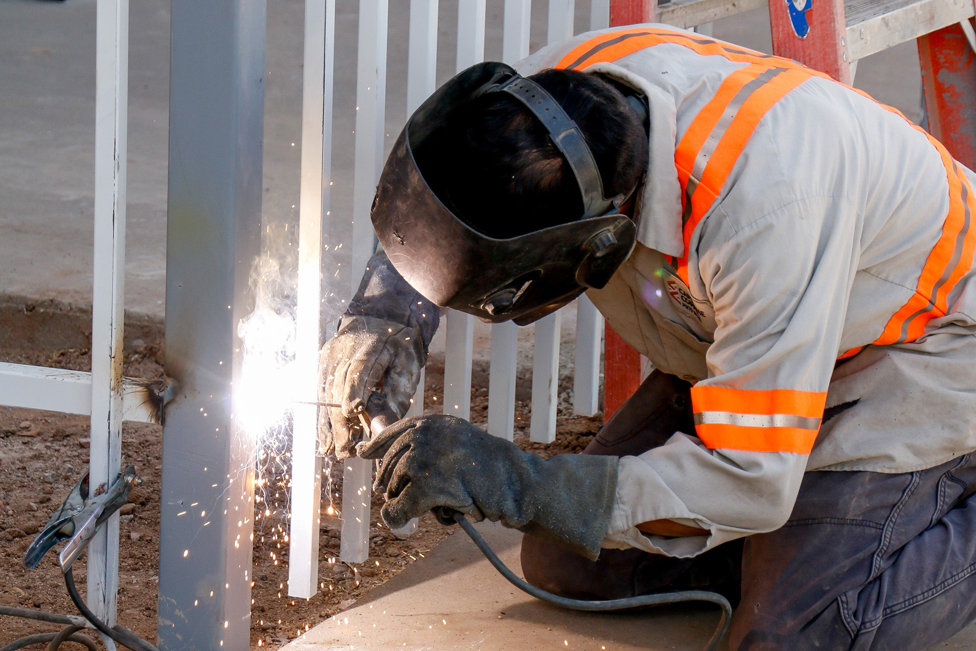 A man in safety equipment kneels as he uses welding tools on a gate