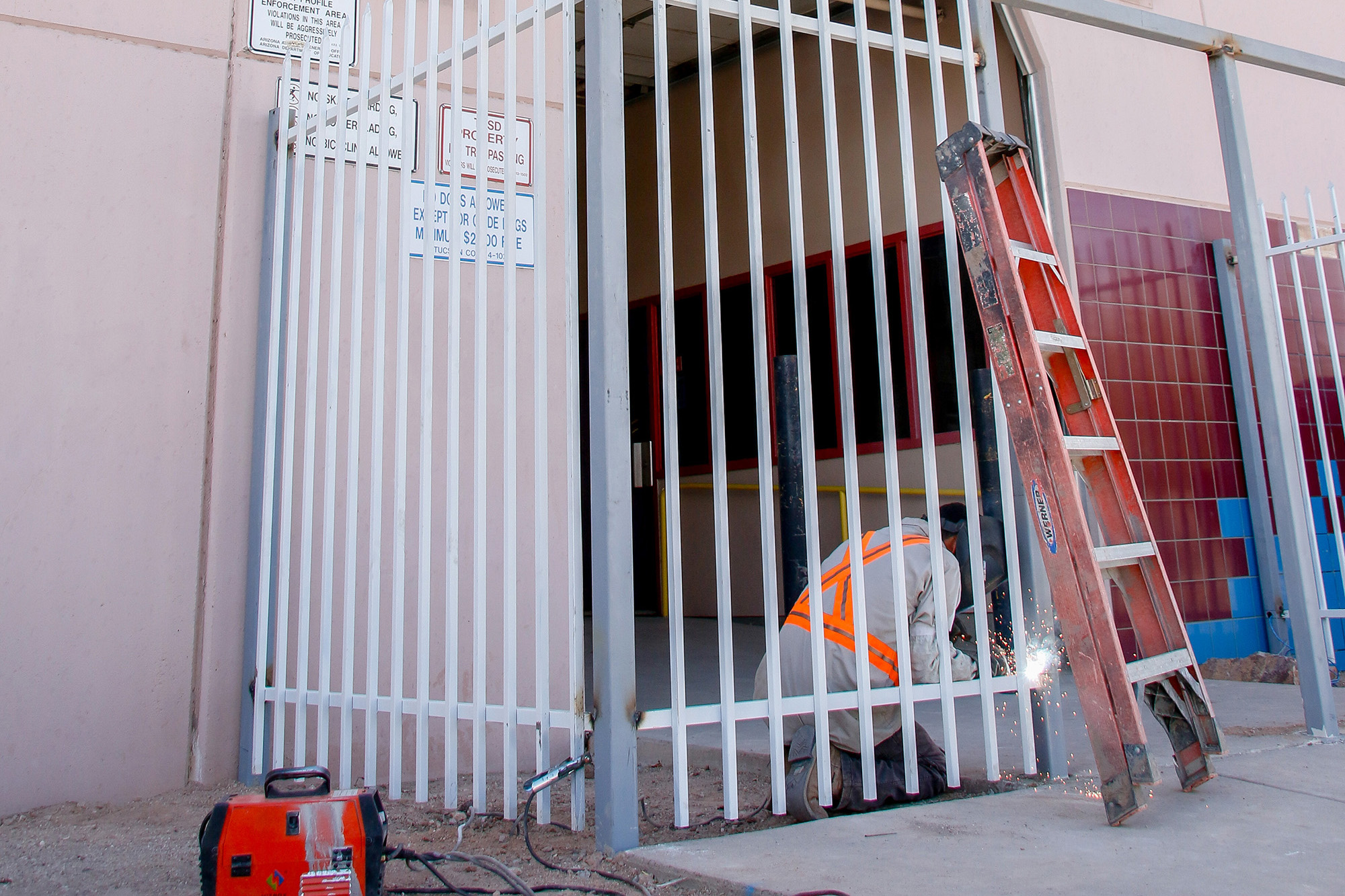 A man kneels on the ground behind a gate using welding tools on the bars